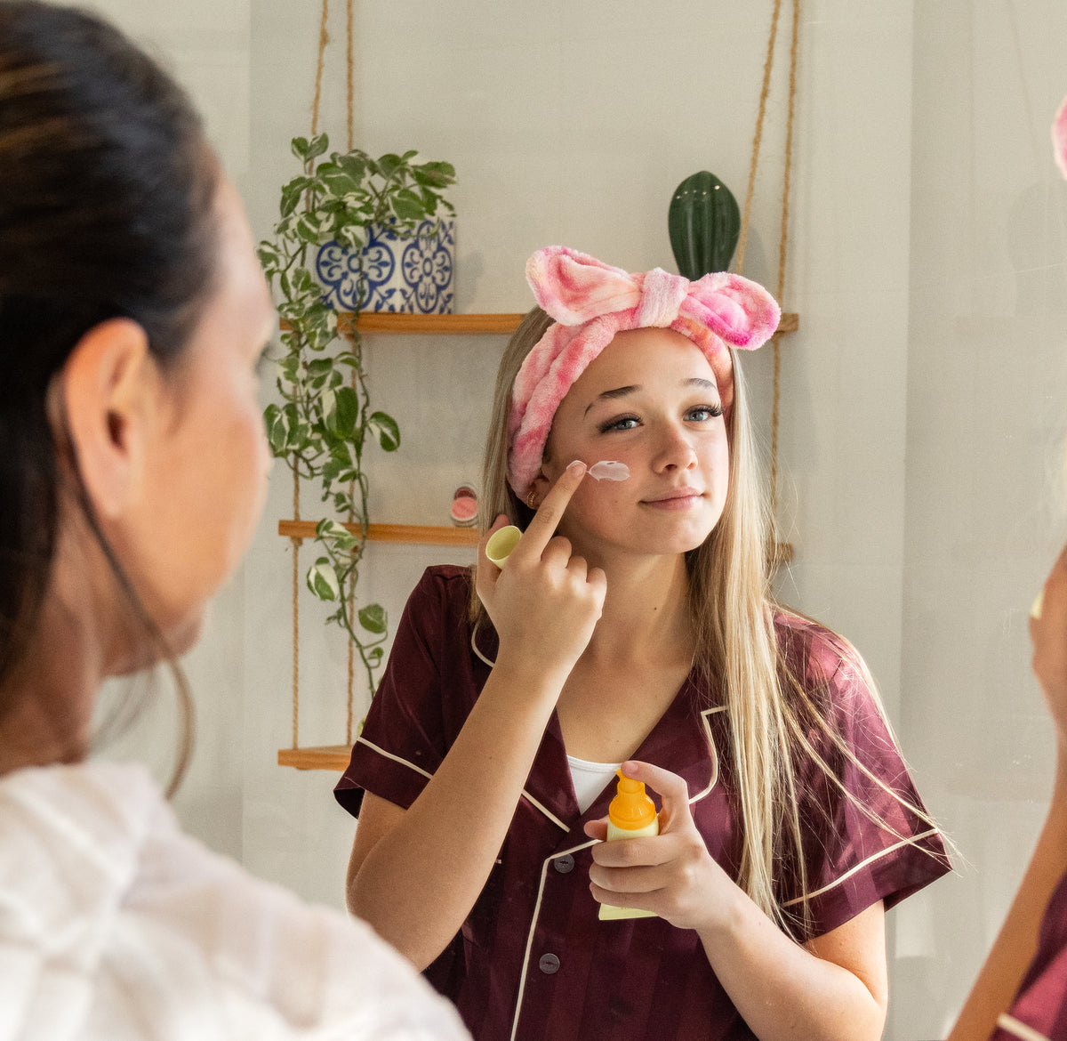 Mother watchin teenage daughter applying cream to face with a pink fluffy tied-dyed headband on her head, in a home setting.